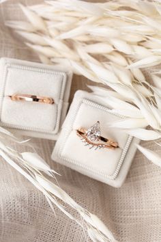 two engagement rings sitting on top of a white cloth covered box next to some dry grass