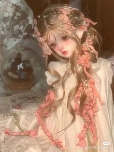 a doll with long hair wearing a white dress and pink flowers in her hair, sitting on a bed