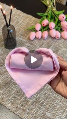 a person holding a pink heart shaped napkin with candles in the background and some flowers