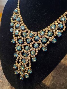 "Our photos do not do justice to this dazzling D and E Juliana bib necklace. The color is more of a gorgeous aqua rivoli stone surrounded by flashes of brilliant gold AB rhinestones threaded throughout this vintage spectacular piece of jewelry from the 60's. A lot of the D and E bib drop necklaces are designed in reds and oranges and deep blues, while this piece fluctuates in the gorgeous aqua and blue and green ranges. That renders this glorious creation the ability to be worn with many more de Vintage Blue Necklace For Evening, Retro Blue Jewelry For Party, Vintage Blue Jewelry For Evening, Blue Vintage Jewelry For Evening, Blue Mid-century Jewelry For Formal Occasions, Mid-century Blue Formal Jewelry, Vintage Blue Rhinestone Jewelry, Vintage Blue Jewelry For Party, Drop Necklaces