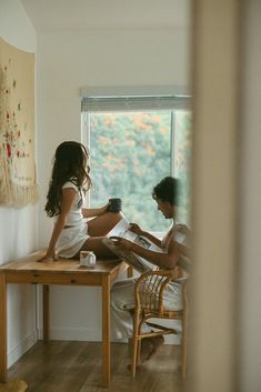 two people sitting at a wooden table in front of a window, one reading a book