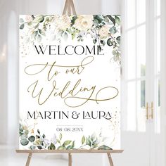 a welcome sign with flowers and greenery in gold foil on an easel next to a window