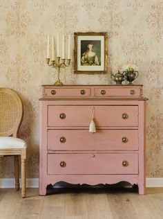 a pink dresser sitting next to a chair in a room with wallpaper and a painting on the wall