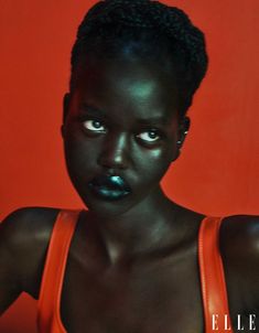 a woman with dark skin and black makeup looks at the camera while wearing an orange tank top