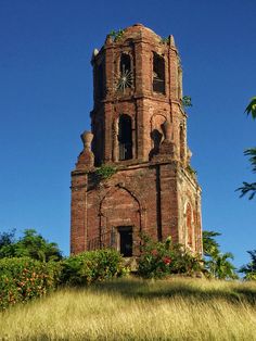 an old brick clock tower sitting on top of a lush green hillside under a blue sky