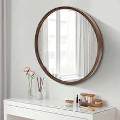 a round mirror on the wall above a white table with makeup and flowers in vases