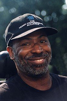 a smiling man wearing a black hat and sitting in a chair with trees in the background