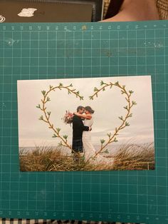 a person holding up a photo in front of a green cutting mat with a heart shaped frame