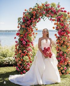 a woman in a wedding dress standing under an archway with flowers on the grass and water behind her