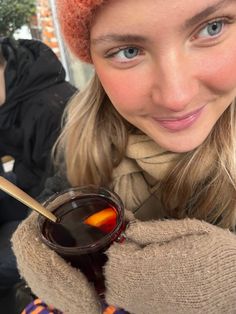 a woman holding a cup of tea with a spoon in it and wearing a knitted hat