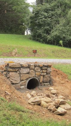 a stone tunnel in the middle of a dirt field with trees and grass behind it