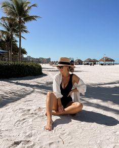 a woman sitting on top of a sandy beach wearing a hat and black bathing suit