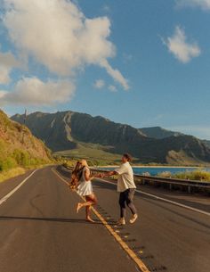 a man and woman walking down the middle of an empty road with mountains in the background