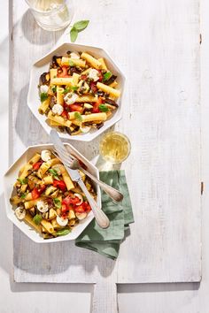 two bowls filled with pasta and vegetables on top of a white wooden table next to a glass of wine