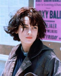 a woman with short hair standing in front of a pink sign and wearing a leather jacket