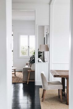 a living room with white walls and black flooring in the center, along with a wooden dining table surrounded by two beige chairs