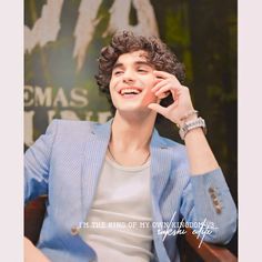 a young man sitting in a chair talking on a cell phone while wearing a blue blazer