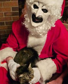 a man dressed as santa claus holding a cat while wearing a white mask and red coat