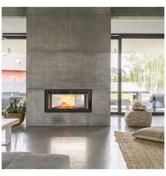 a living room with concrete walls and a fire place in the center, surrounded by large windows