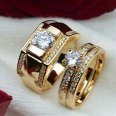 two gold wedding rings sitting on top of each other next to a red rose and white cloth