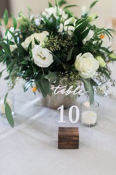 a table topped with a vase filled with white flowers and greenery next to candles