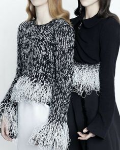 two models in black and white clothing with long sleeves, one wearing a sweater that has fringes on it