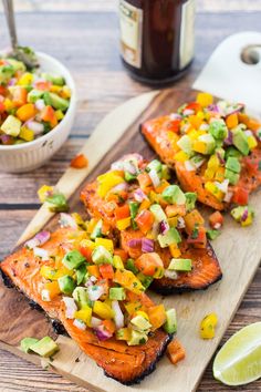 grilled salmon with mango salsa and avocado on a wooden cutting board next to lime wedges