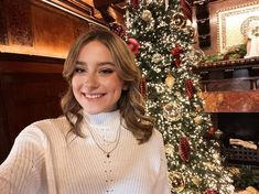 a woman standing in front of a christmas tree wearing a white sweater and smiling at the camera