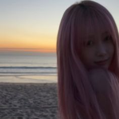 a woman with long pink hair standing on the beach at sunset, looking off into the distance