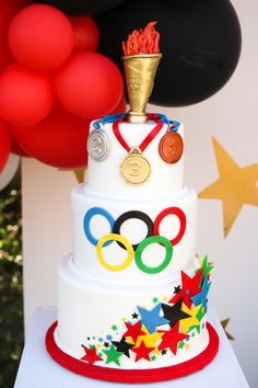 a three tiered cake with olympic symbols on it and red balloons in the background