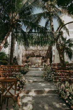 an outdoor ceremony setup with wooden chairs and greenery on the aisle, surrounded by palm trees