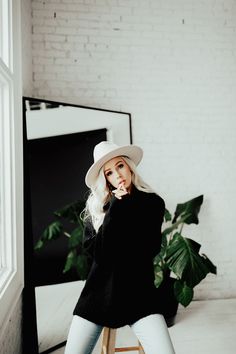 a woman sitting on top of a wooden stool next to a window wearing a white hat