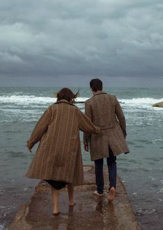 two people are walking along the edge of a pier towards the ocean on a cloudy day