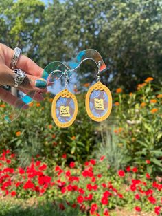 a person holding up some earrings in front of red flowers and green grass with trees behind them