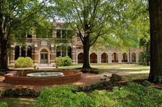 an old brick building surrounded by trees and shrubbery with a fountain in the foreground