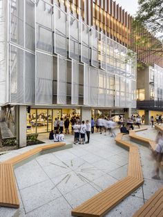 people walking and sitting on benches in front of a large building with glass walls,