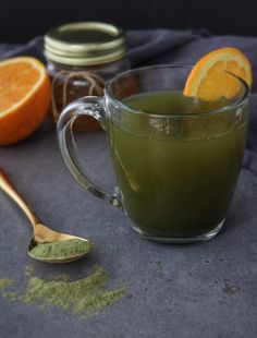 a glass mug filled with green liquid next to an orange slice and spoon on a table