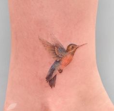 a small hummingbird tattoo on the side of a woman's foot, with an orange and blue tail