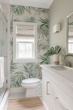 a white toilet sitting under a bathroom window next to a sink and bathtub covered in green palm leaf wallpaper