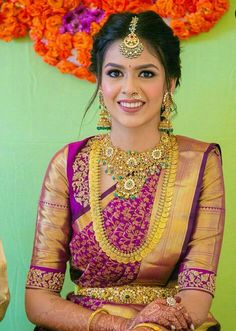 a woman in a purple and gold sari sitting on a chair with flowers behind her