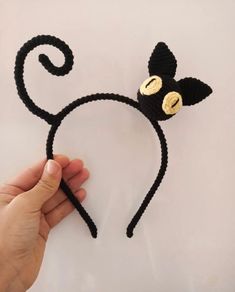 a hand is holding a crocheted black cat headband with two gold eyes