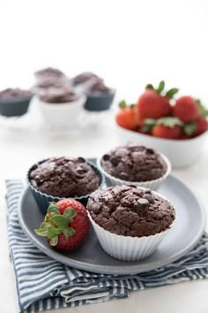 three chocolate muffins on a plate with strawberries and other dessert items in the background