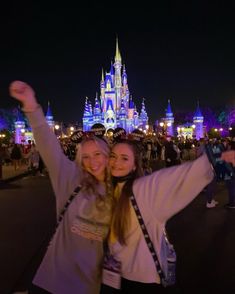 two women taking a selfie in front of the disney castle at night with their arms up