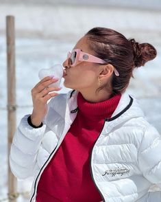 a woman in white jacket and pink sunglasses blowing bubbles into her mouth while standing on snow covered ground