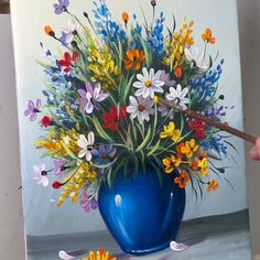 a painting of colorful flowers in a blue vase on a white surface with a person's hand holding a paintbrush