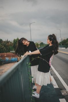 two young women standing on the side of a road next to a green metal fence