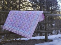 a pink and white quilt hanging on a wooden fence next to snow covered ground with trees in the background