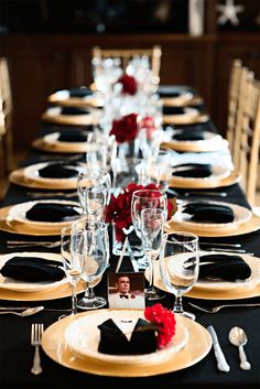 a table set with black and gold plates, silverware and red flowers on it