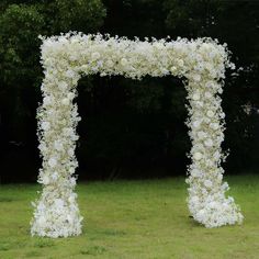 a large white flower covered archway in the middle of a grass field with trees behind it