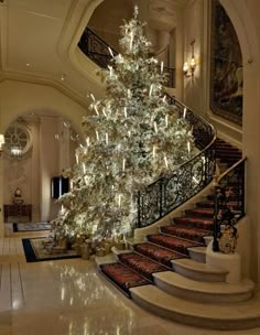 a large christmas tree in the middle of a room with stairs and chandelier